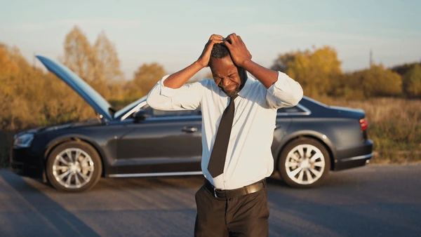 rental-car-breakdown | Rwanda Car Rental Services Man in a white shirt and black tie stands with hands on his head in front of a dark sedan with its hood open on a rural road appearing frustrated by a breakdown