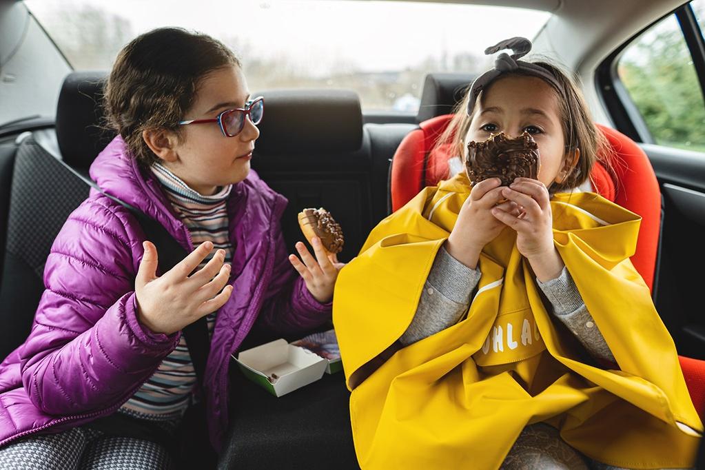 Generated by pixel 2026-08-07T061411893636 | Rwanda Car Rental Services Two young girls sit in a cars backseat eating chocolate donuts one in a purple jacket and glasses the other in a yellow cape